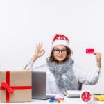 Une femme portant un bonnet de Noël fait un signe « OK » tout en tenant une carte de crédit à un bureau avec un ordinateur et des cadeaux, illustrant des astuces pour des achats de Noël sans stress.