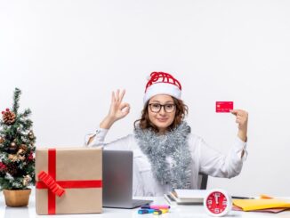 Une femme portant un bonnet de Noël fait un signe « OK » tout en tenant une carte de crédit à un bureau avec un ordinateur et des cadeaux, illustrant des astuces pour des achats de Noël sans stress.